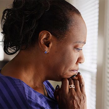 Gloria looking worried, staring out the window of her home through the blinds